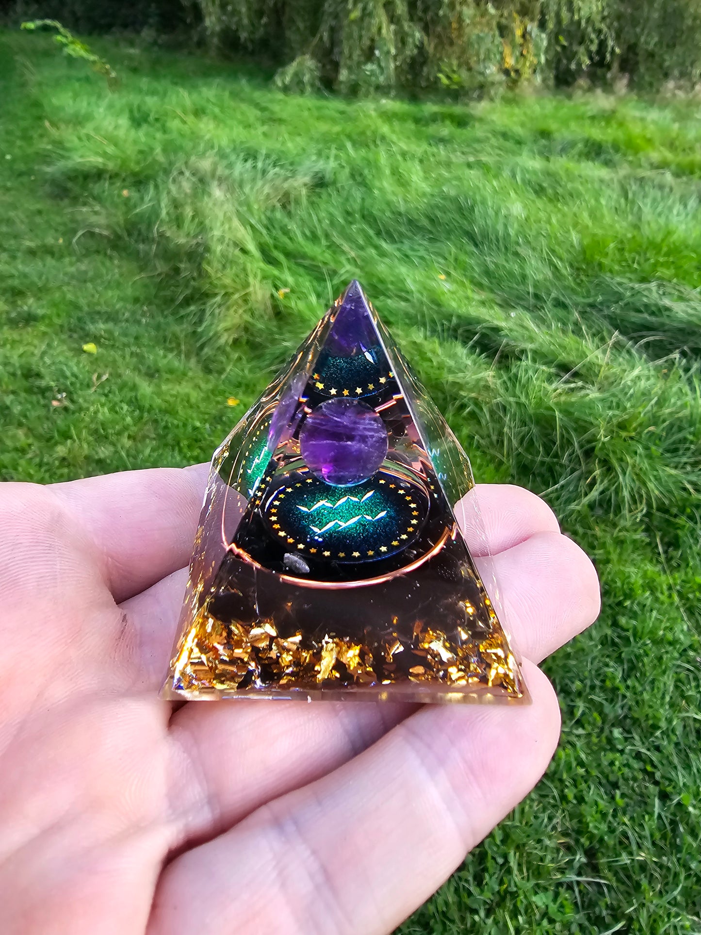 Håndlavet orgonit pyramide med ametyst kugle, stjernetegn og gyldne flager, symboliserer harmoni og balance, mod en naturlig baggrund. Fra Naturens Skatte.