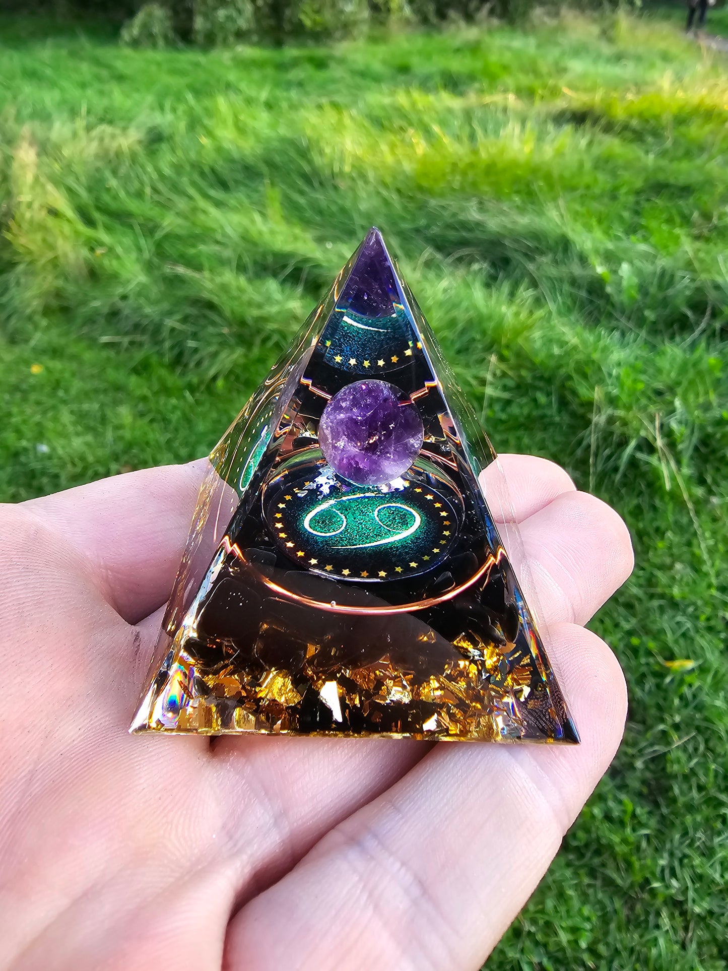 Håndlavet orgonit pyramide med ametyst kugle, stjernetegn og gyldne flager, symboliserer harmoni og balance, mod en naturlig baggrund. Fra Naturens Skatte.